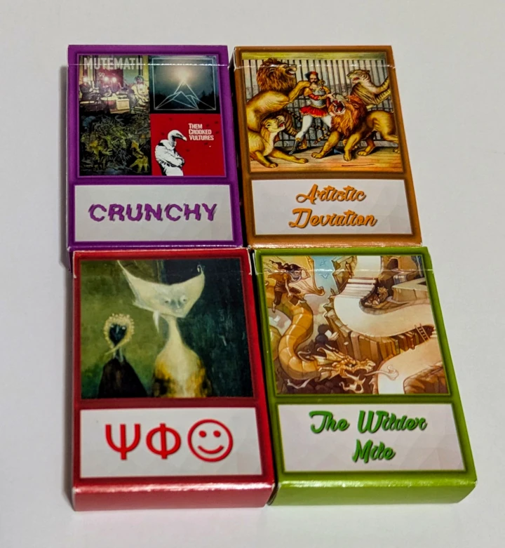 The fronts of four deck boxes, showing paintings or album covers, and the deck names (Crunchy, Artistic Deviation, The Wilder Mile, and some greek symbols)